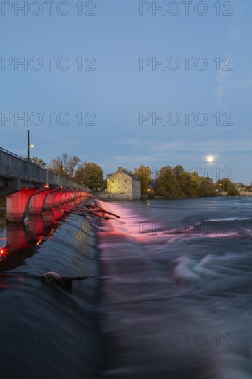 Illuminated Moulin Neuf water flow control dam and walkway over Des Mille-Iles river plus New Mill on Ile des Moulins in autumn at dusk, Old Terrebonne, Quebec, Canada