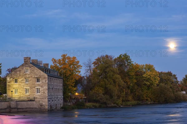 Illuminated Moulin Neuf - New Mill on Ile des Moulins and Des Mille-Iles river with supermoon rising in autumn at dusk, Old Terrebonne, Quebec, Canada
