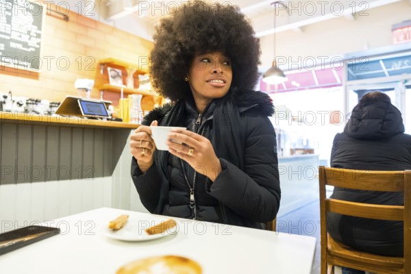 Young african american woman relaxing in a cozy manhattan coffee shop, enjoying a hot coffee during winter, embodying urban lifestyle, leisure, and social connection