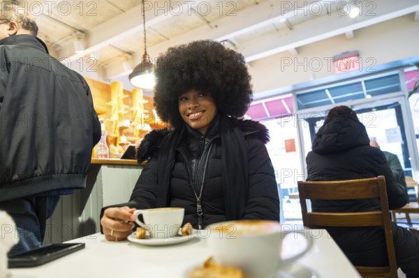Young black woman with an afro hairstyle smiling while holding a cup of coffee at a table in a bustling cafe, representing urban lifestyle, casual leisure, and diversity in new york city