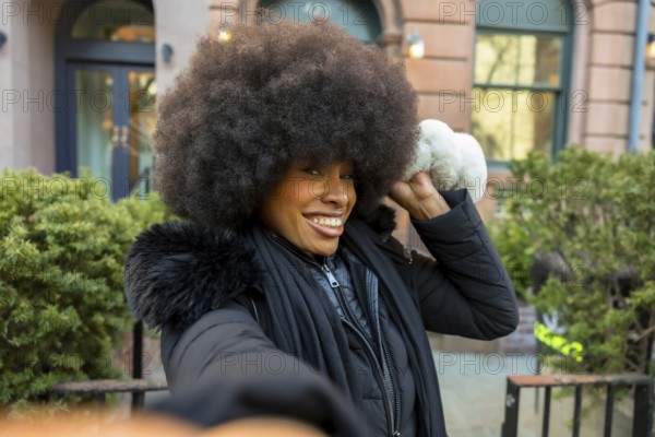African american woman with afro hairstyle smiling and preparing to throw an artificial snowball during winter in manhattan, embodying playful seasonal joy