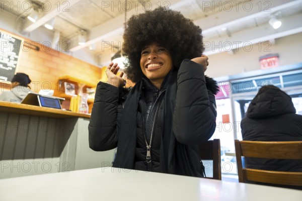 Smiling black woman with a large afro in a warm black puffer jacket sitting in a casual manhattan cafe, laughing and enjoying a cozy, confident moment over coffee