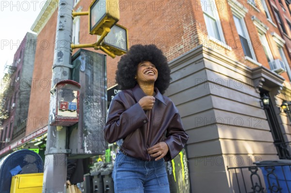 Young african american woman with afro hair smiling and looking up, walking in a vibrant new york city neighborhood street with a brick building and urban infrastructure