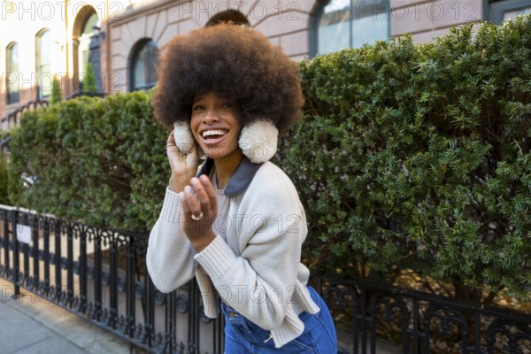 Young african american woman with an afro hairstyle laughing brightly, wearing warm earmuffs and a sweater while enjoying the vibrant street atmosphere in manhattan during winter