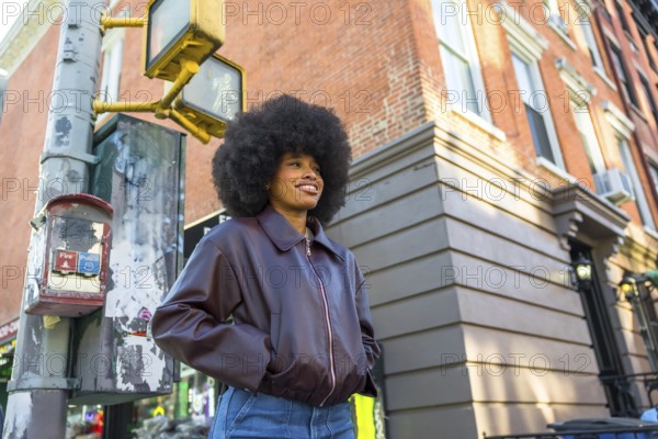 Young woman smiling while standing confidently on a new york city street corner, featuring an afro hairstyle and casual fashion with old brick architecture in the background