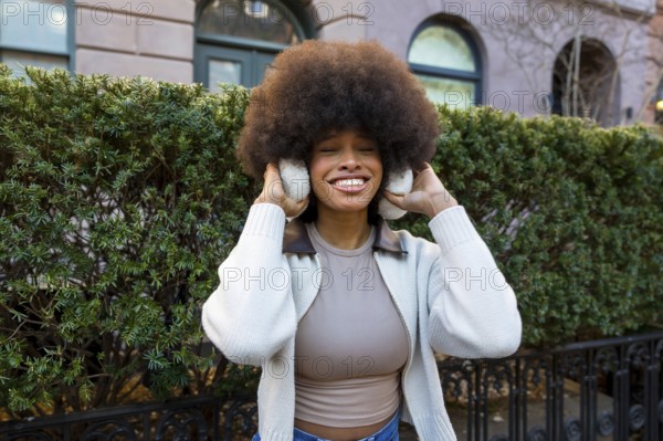 Smiling black woman with large afro hair wearing earmuffs and a sweater, standing outdoors on a winter day in new york city with a green hedge behind her