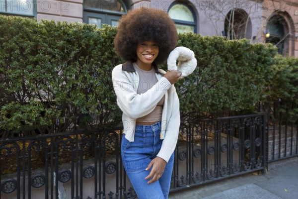 African american woman smiling, showing confident joy and modern fashion, holding a unique fluffy handbag while standing on a new york city street with green hedges and classic architecture