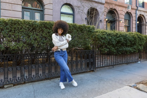 Young black woman with an afro hairstyle smiling confidently while standing on a sidewalk in front of residential buildings, embodying urban lifestyle, joy, and diversity in a city environment
