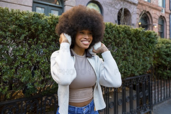 African american woman wearing earmuffs and a sweater, smiling brightly while looking away in a vibrant neighborhood of new york city, enjoying urban fashion and natural beauty