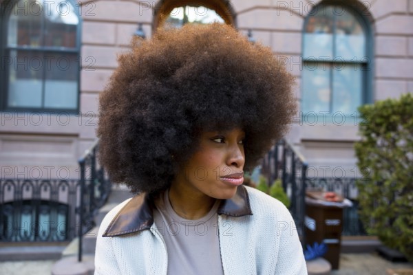 African american woman with a large afro hairstyle looking away, wearing a casual jacket, standing outdoors in front of a manhattan brownstone building representing urban life