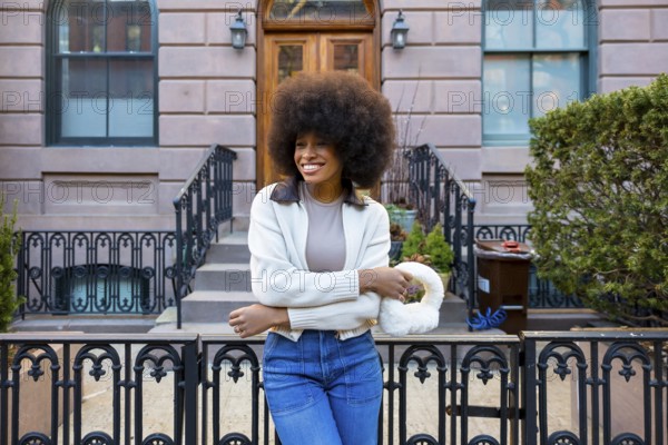 Smiling young woman with a large afro hairstyle and fashionable attire standing in front of a traditional brownstone building in new york city, enjoying urban life