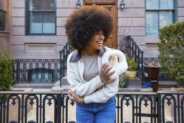 Happy black woman with afro hair laughing, holding furry earmuffs, and wearing jeans with a sweater in an urban setting during cold weather, representing joy, style, and authenticity