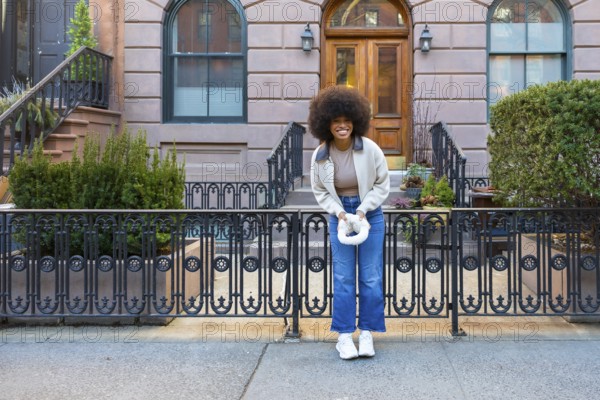 Smiling young black woman with afro hairstyle standing happily on a sidewalk in front of a classic brownstone apartment building, embracing urban lifestyle and positive vibes