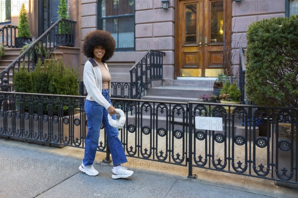 Happy young woman with an afro hairstyle walking on a sidewalk in front of brownstone buildings in manhattan, new york city, wearing casual streetwear and carrying a modern handbag