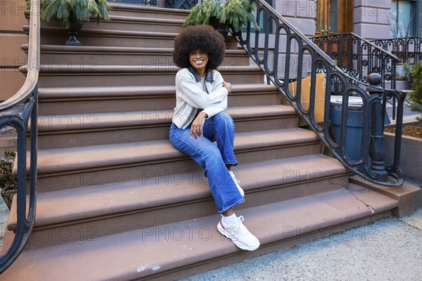 Young african american woman with afro hair sitting on a brownstone stoop in a new york city neighborhood, enjoying urban lifestyle and modern casual fashion