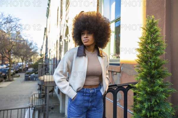 Young woman with an impressive afro hairstyle and casual fashion standing on a residential street during golden hour, reflecting urban lifestyle, confidence, and individuality