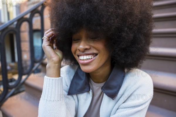 Young black woman laughing with eyes closed and hand in hair, sitting on a brownstone stoop, showcasing joy and spontaneity in an urban new york setting