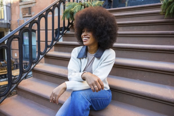 Young black woman with afro sitting on harlem brownstone stoop, smiling and relaxed, enjoying sunny nyc street life confident, stylish, free and authentic urban joy