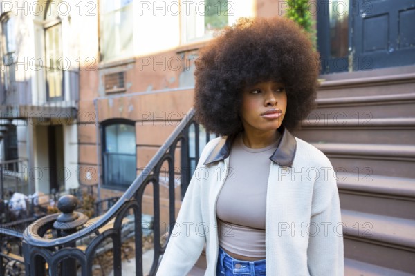 Young black woman with natural afro hair standing on an urban brownstone stoop, looking away while portraying concepts of individuality, urban life, and street style in manhattan