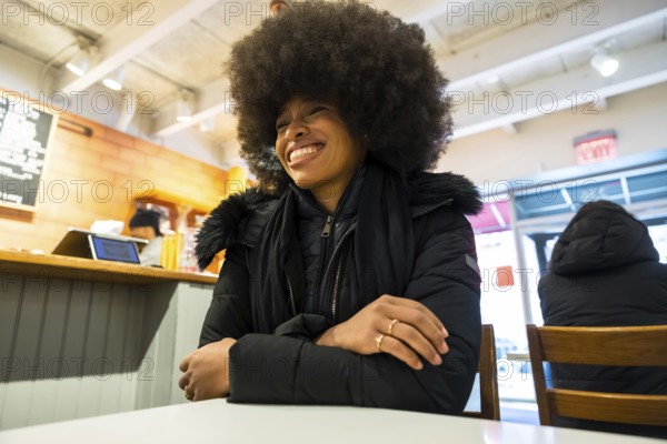 Smiling black woman in a winter jacket chatting happily inside a bustling manhattan coffee shop, enjoying a cozy, relaxed moment of urban comfort and confident contentment