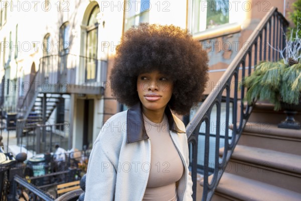 Young african american woman with voluminous afro hair standing on the steps of a brownstone building in new york city, looking away, reflecting urban lifestyle and beauty