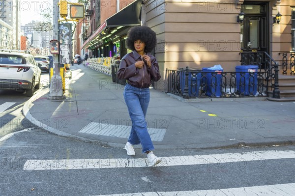 Young woman with afro hair and casual fashion attire confidently walking across a zebra crossing in a vibrant new york city urban street, representing city life and modern style