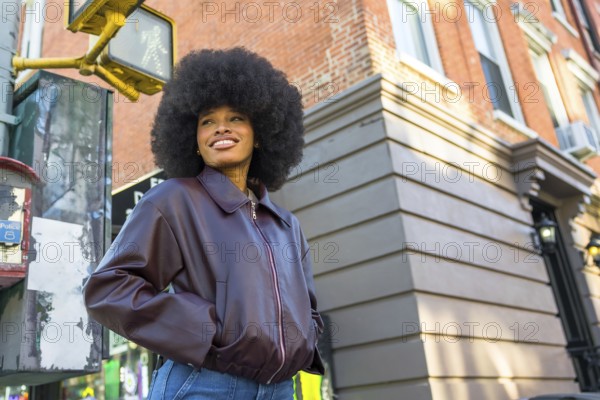 Young african american woman wearing a leather jacket and jeans, standing at a city intersection in manhattan, smiling and looking away while enjoying the vibrant street scene
