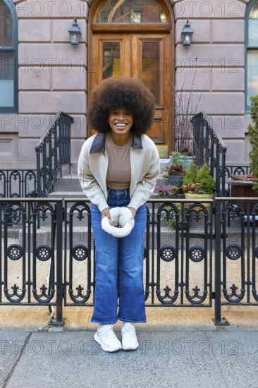 Smiling woman with afro hair holding a white furry hand warmer, standing on a sidewalk in front of a classic new york city brownstone building with an ornate gate