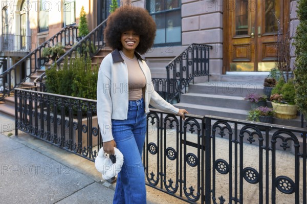 Young black woman with a large afro in jeans and a sweater, smiling confidently while standing on a manhattan brownstone sidewalk, enjoying sunny urban street style