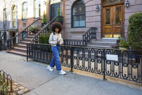 Young woman with afro hair walking down a sidewalk in a historical new york city neighborhood, holding a fashionable accessory and exploring the urban environment