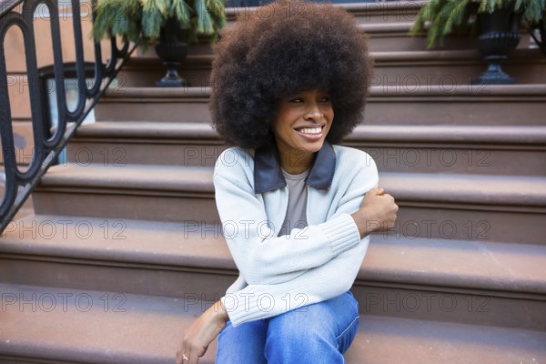 Young black woman with an afro hairstyle smiling brightly while sitting on the brownstone steps of a building, expressing joy and urban lifestyle in new york city