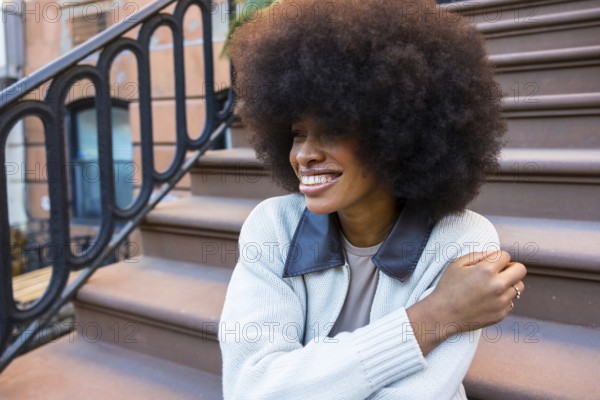 African american woman with a beautiful afro hairstyle sitting and smiling on brownstone steps in manhattan, embodying urban lifestyle, joy, and individual expression