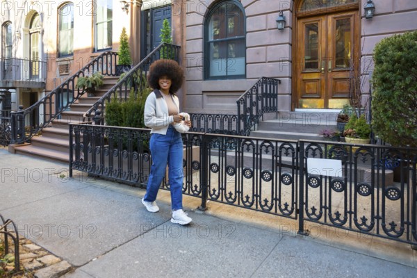 Young african american woman with afro hair smiling while walking along a new york city sidewalk past classic brownstone residential buildings, representing urban lifestyle and individuality