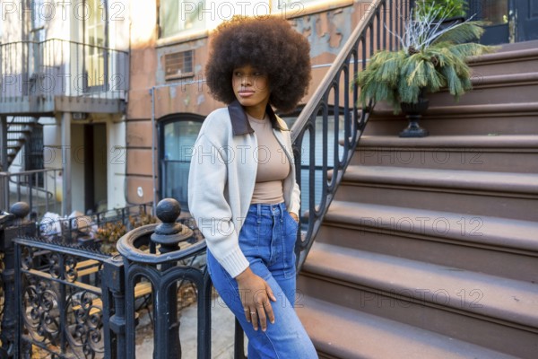 Stylish young woman with afro hair posing confidently on the brownstone steps of an urban building, embodying modern street fashion and a vibrant city lifestyle in harlem, new york