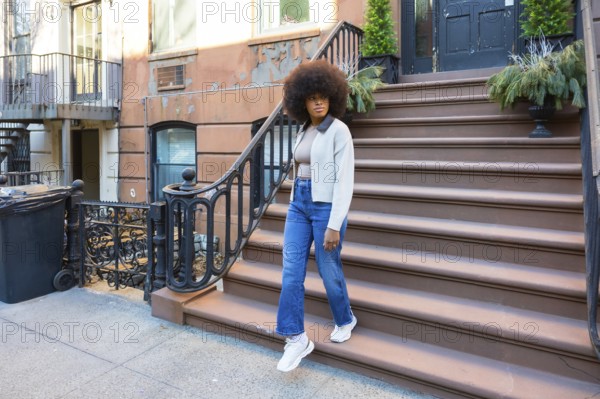 Young black woman with a stylish afro hairstyle walking down the steps of a traditional brownstone building, expressing urban living, fashion, and independence in a vibrant city setting