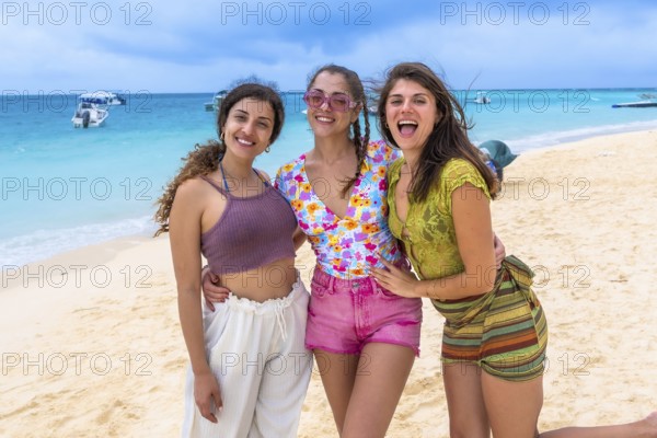 Three happy young women standing together on a white sandy beach, smiling and having fun during their tropical summer holidays with turquoise sea in the backdrop