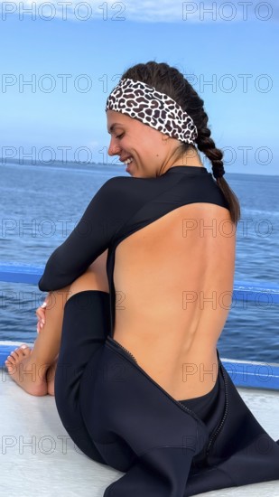 Woman wearing a wetsuit and leopard print headband, smiling happily while sitting on a boat deck, preparing for watersports activities in the clear blue sea of the maldives
