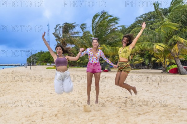 Three happy young women friends jumping while holding hands on a sandy beach, celebrating freedom and friendship during a tropical vacation to the maldives