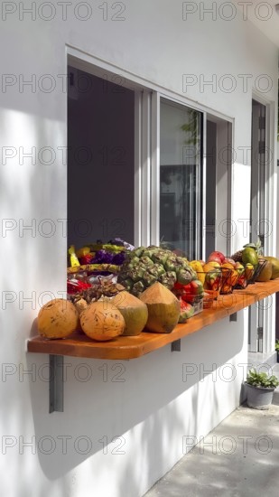 Assortment of fresh tropical fruit and young raw coconuts displayed on a wooden shelf outside a building window, offering healthy refreshments in a sunny outdoor setting