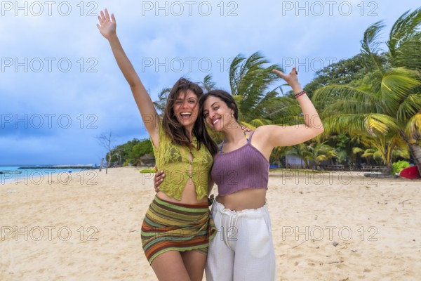 Two smiling young women friends embracing, raising arms, and expressing joy, celebrating their fun tropical beach holiday with palm trees swaying in the background