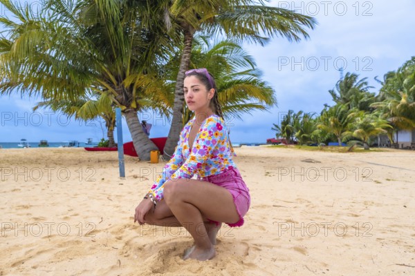Confident young woman wearing colorful summer clothes posing on a sandy tropical beach surrounded by palm trees under a cloudy sky during a relaxing holiday in the maldives