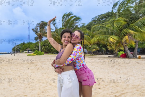 Two happy young women friends embracing on a tropical sandy beach with palm trees and a clear blue sky, enjoying their summer island vacation in the maldives