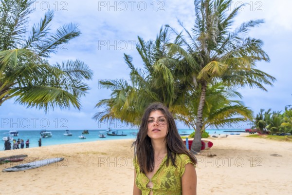 Woman standing on a tropical beach in the maldives, surrounded by sand, palm trees, and turquoise ocean water with boats visible in the background under a cloudy sky