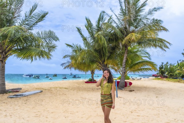 Woman smiling and standing on a beautiful sandy beach fringed with green palm trees and turquoise ocean water with boats in the background, representing travel and leisure