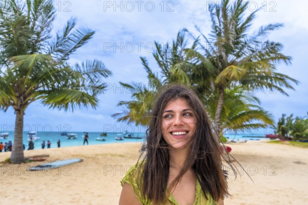 Young woman smiling and looking away, standing on a sandy beach with palm trees and a turquoise ocean bustling with boats in the background, embodying travel, freedom, and relaxation