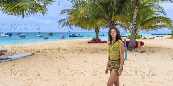 Woman stands on sunlit sandy beach under palm trees, wearing green top and sarong while gazing at turquoise lagoon with anchored boats and distant island horizon