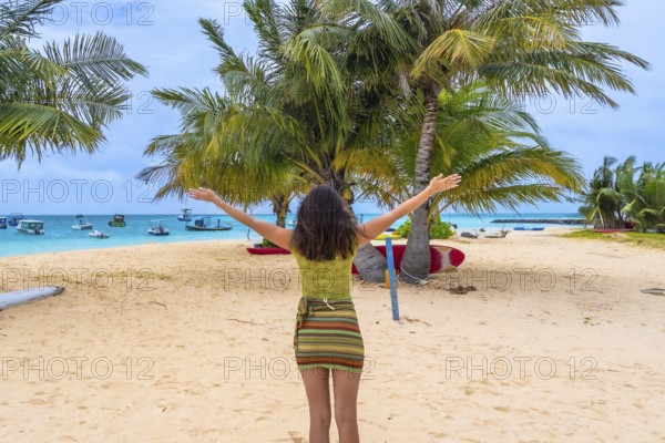 Woman standing on a tropical sandy beach with arms outstretched, experiencing a joyful moment of freedom and relaxation amidst lush palm trees and turquoise ocean waters with boats in the background