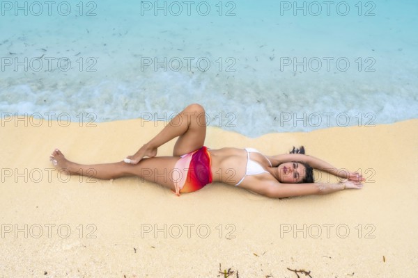 Young woman lying on the pristine sandy beach by clear turquoise ocean water, enjoying a tropical vacation getaway in the sun, finding peace and relaxation