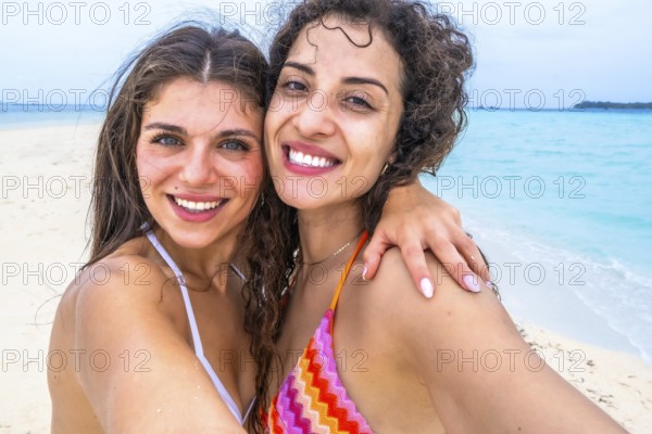 Young women smiling for a selfie on a sandy beach, embracing during a tropical island vacation, enjoying friendship and travel in a sunny ocean destination
