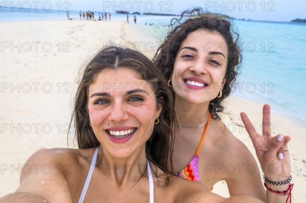 Smiling women capturing a joyful selfie on a beautiful white sand beach with turquoise water during a tropical vacation to the maldives, symbolizing friendship and travel memories